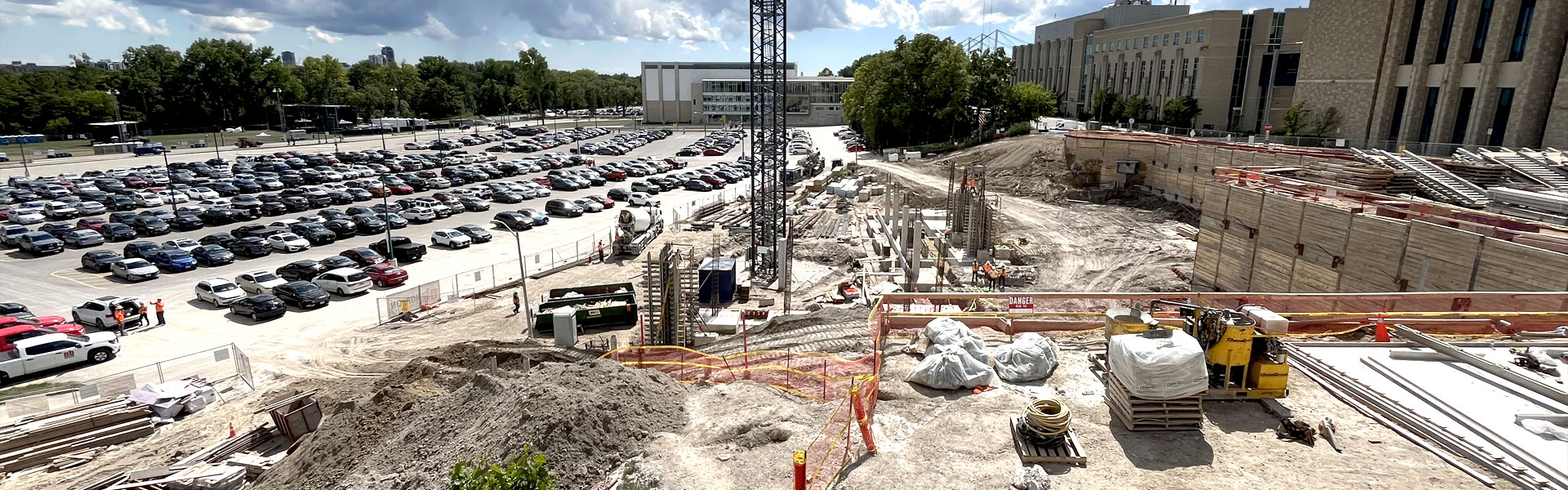Image of the construction site at engineering with a retaining wall to the right and the south valley parking lot to the left. The base of crane sits in the middle of the site and workers in high vis shirts are busy working on various things.
