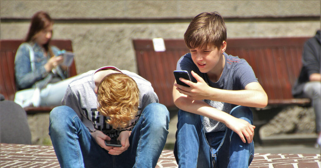 Two boys sit on the steps outdoors. Both are on their phones.